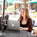 A female student sits in front of her laptop. She has long blond hair and pale skin. She is smiling as she looks off into the distance. 