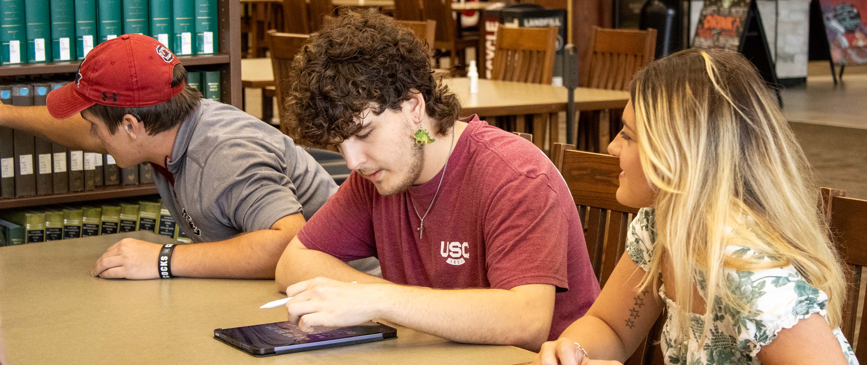 At Thomas Cooper Library, a student picks a book off the shelf while another uses their tablet.