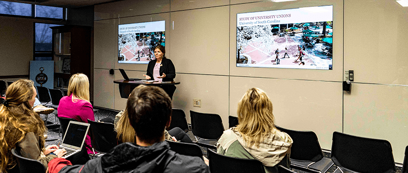 An audience watches a presenter in front of tech walls. 