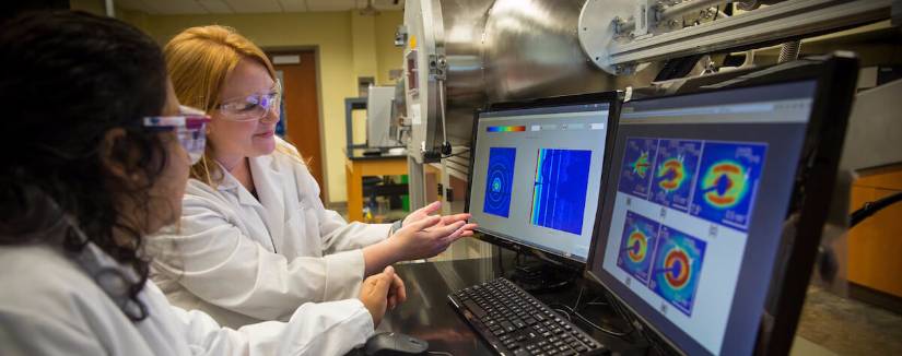 Student and professor in white lab coats and safety classes work together in a lab, examining images on two computer screens