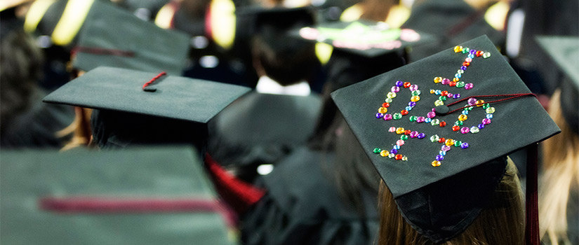 View of the top of multiple mortarboards at graduation, including one decorated in multicolored rhinestones to spell out the phrase I Did It