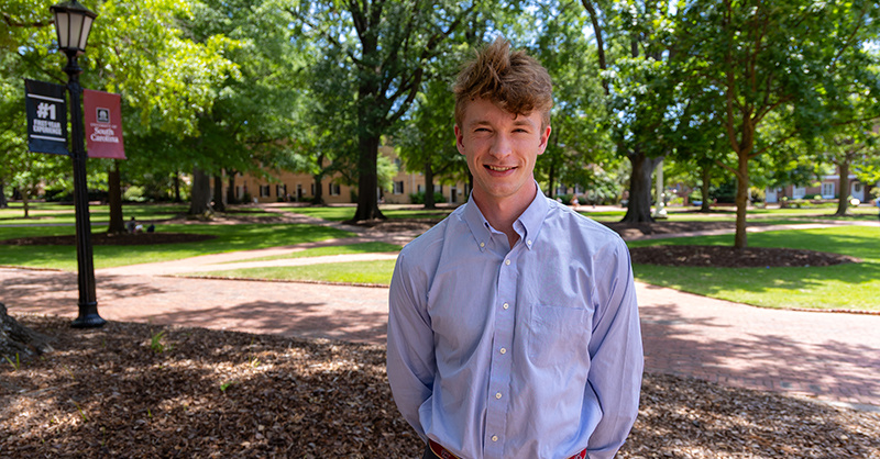Christian Geils stands smiling on the University of South Carolina’s historic Horseshoe, wearing a light blue button-down shirt, with trees, brick walkways, and a campus sign in the background.