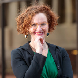 Rebecca has red hair and is wearing a black blazer, green blouse, and geometric earrings. She stands in front of a brick building