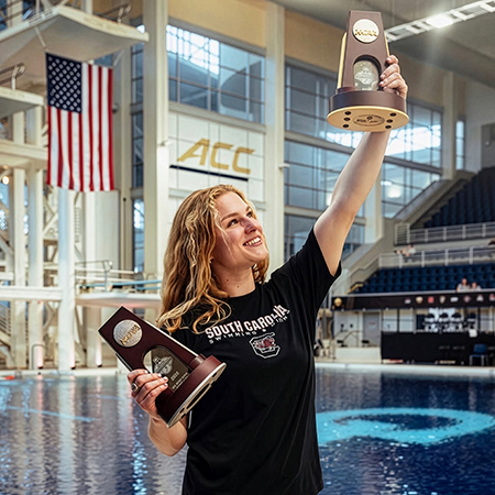 Sophie Verzyl raising NCAA trophy poolside with ACC signage and American flag in background