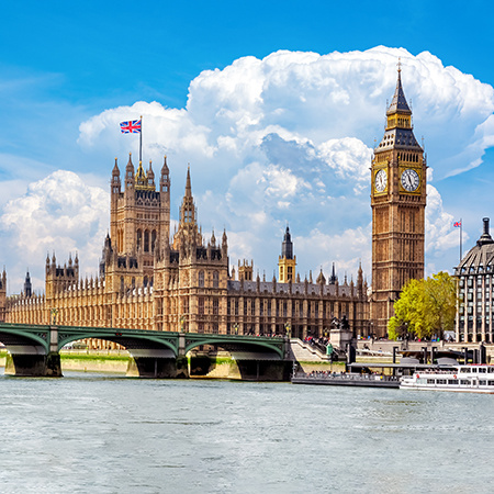 The Palace of Westminster and Big Ben tower along the River Thames in London, with a Union Jack flag flying under a bright cloudy sky.
