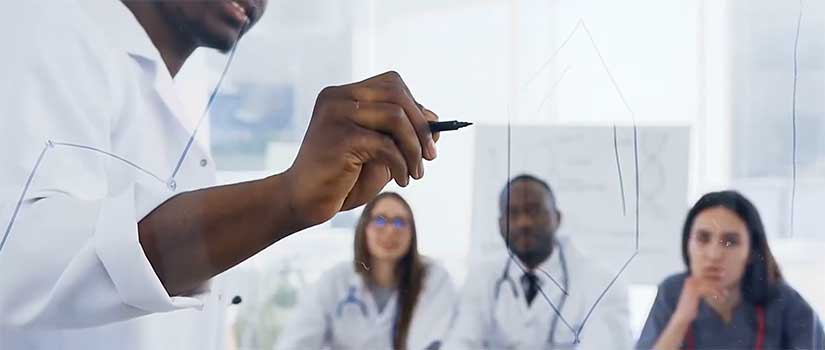 Medical student in white coat writes a formula on clear glass while other watch.