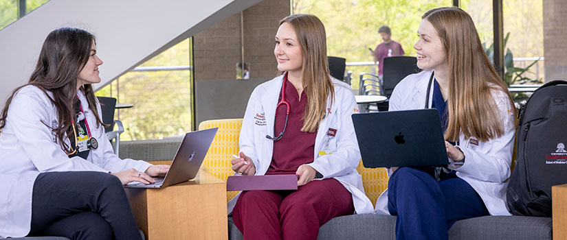 Male med student in white coats in sim lab. One school can better the world logo.