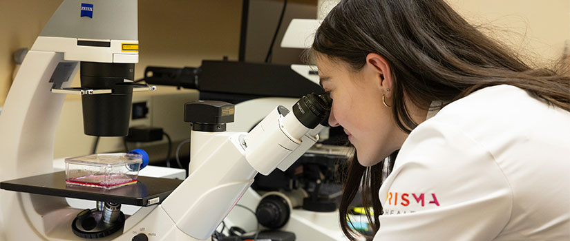 Student in lab coat peers into microscope. 