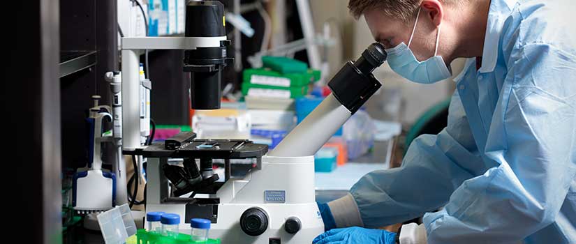 Student in blue labcoat and mask peers into microscope in lab.