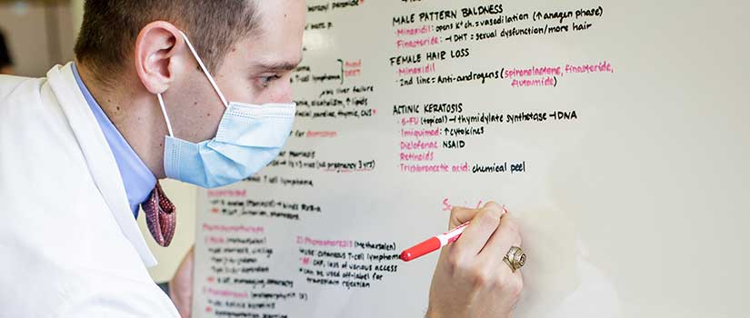 Student in white coat and mask writes on whiteboard.