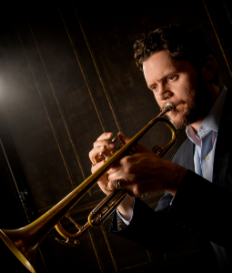 male dressed in black suit playing the trumpet with black background