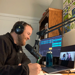 Visual communications instructor Jason Porter wears headphones and sits in front of a microphone and laptop